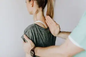 Back view of crop unrecognizable osteopath in uniform and wristwatch checking up back of slim female patient in casual wear on white background